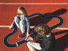 Two children play with the Large Flexible Toy Race Track - Grand Prix, set up on a red outdoor surface with white lines. One child wears grey, and the other wears a black shirt with white dots.