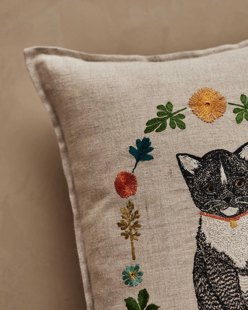 The Coral & Tusk Kitten Cameo Pillow features a beige linen cover with embroidered flowers and a partial black and white cat in a red collar, making it perfect for springtime decor on any neutral background.