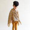 A young child stands against a plain background, looking down while wearing mustard leggings, a beige sweater, and Sarah's Silks Animal Print Playsilk - Cheetah draped over one shoulder.