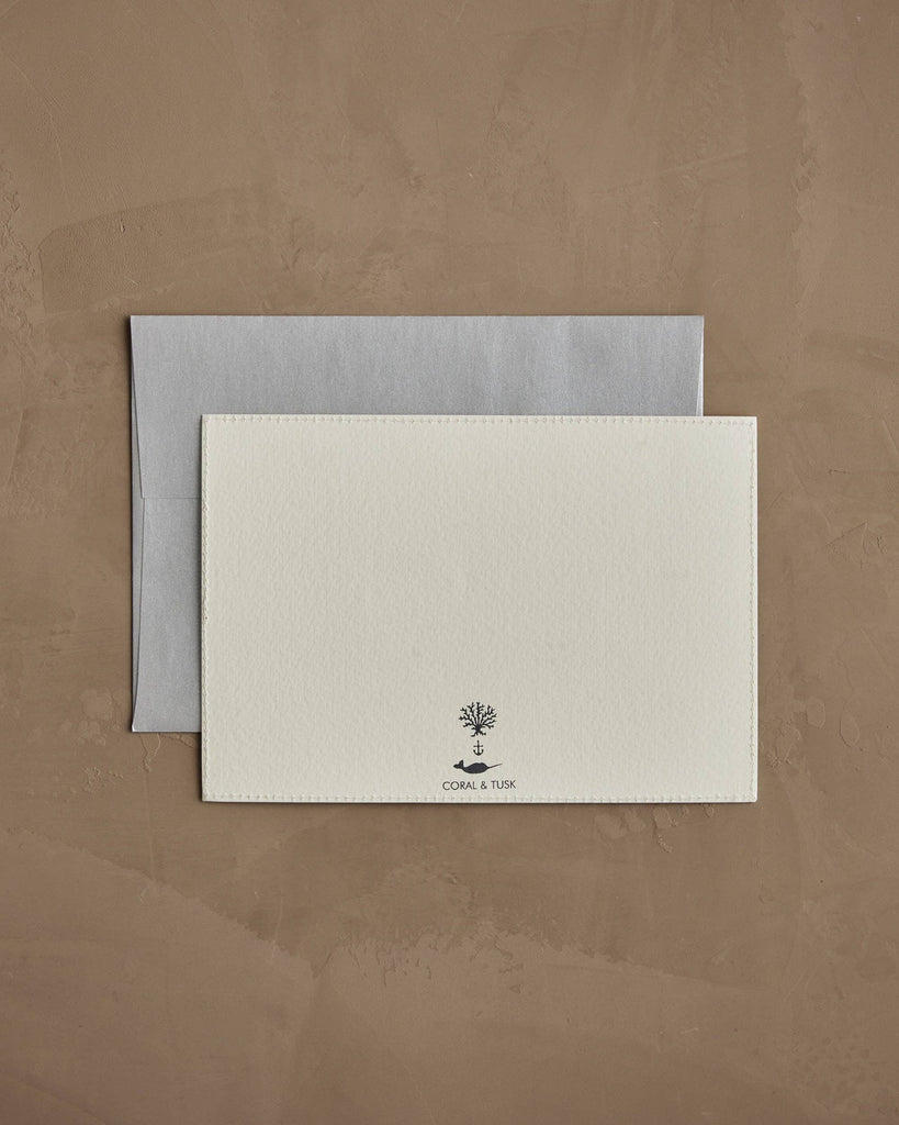 The Coral & Tusk Snowman Card features a blank white card with a tree and anchor logo above "CORAL & TUSK," set on a light gray envelope atop a brown textured surface—perfect for season’s greetings or winter decor.