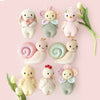 The Cuddle + Kind Baby Chicken, an heirloom-quality handmade doll, is displayed on a pale pink background in two rows with other baby animals and flower dolls, accented by white tulips.