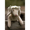 The Senger Naturwelt Stuffed Animal - Small Sheep is a sustainable plush toy featuring a daisy flower crown, resting on a wooden plank with a ladybug on its head and blurred greenery in the background.