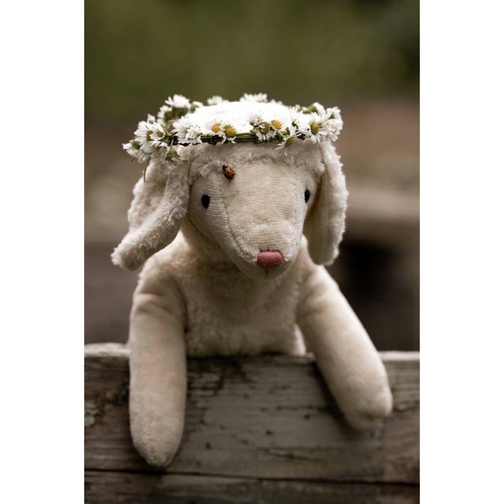 The Senger Naturwelt Stuffed Animal - Small Sheep is a sustainable plush toy featuring a daisy flower crown, resting on a wooden plank with a ladybug on its head and blurred greenery in the background.