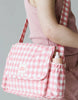 A person carries a stylish Tiny Tummies Pink Gift Box - The Essentials from Tiny Harlow, featuring a pink and white checkered design with a side pocket for doll accessories like a toy bottle. The bag comes with an iconic flap adorned with the "tiny harlow" label. They are wearing a pink textured sleeveless top paired with matching pants.