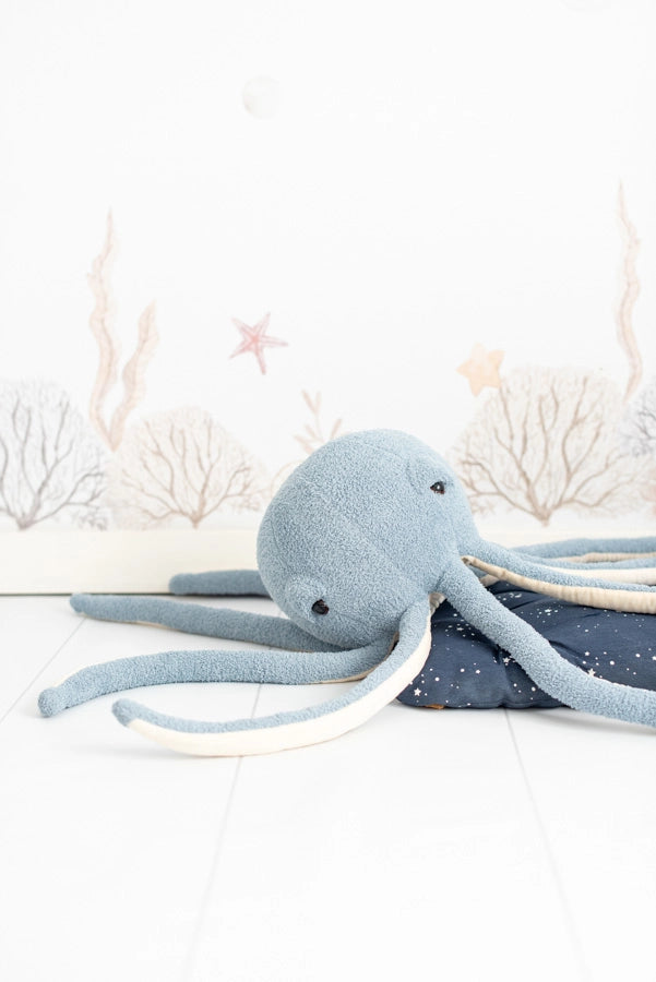 A soft, blue Octopus Stuffed Animal sits on a lightly patterned surface with illustrations of seaweed and starfish in the background, creating a serene underwater theme.