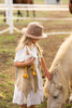 A young girl in a beige hat and white dress stands in a grassy area, holding her favorite Charlie the Chicken Stuffed Animal. She gently pets a small white pony's mane with her other hand. A fenced paddock is visible in the background.