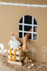 A child reaches through the window of a cardboard house, grabbing eggs from a wire basket. Next to the window, on a small wooden crate, sits Charlie the Chicken Stuffed Animal, with its long yellow legs. The house is decorated with white floral patterns on a brown background.