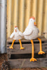Two plush chickens with white bodies and long yellow legs, including the Charlie the Chicken Rattle Stuffed Animal, sit on a wooden crate. One is larger than the other. A pinecone rests nearby against a yellow corrugated metal wall.