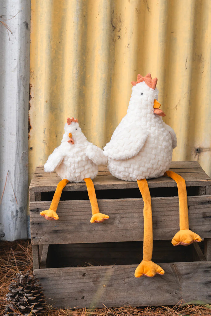 Two plush chickens with white bodies and long yellow legs, including the Charlie the Chicken Rattle Stuffed Animal, sit on a wooden crate. One is larger than the other. A pinecone rests nearby against a yellow corrugated metal wall.