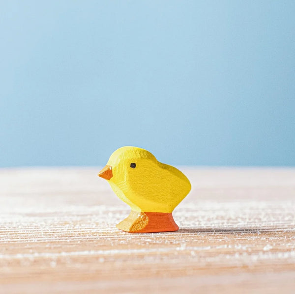 The Bumbu Handmade Wooden Chick, a small yellow figure with an orange beak and feet, stands on a light wooden surface against a soft blue background, highlighting the charm of sustainable wood toys.