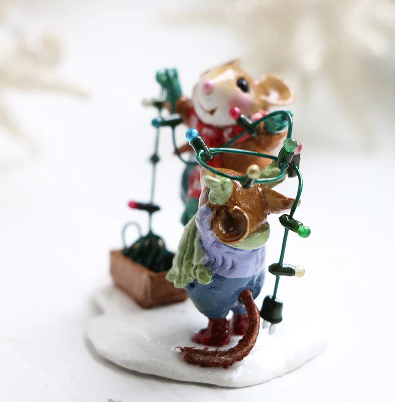 The Wee Forest Folk Tiny Collectible - Merry & Bright features two festive handmade mouse figurines on snow, untangling colorful string lights. One wears a green scarf, the other a red vest, with a brown box of lights beside them.