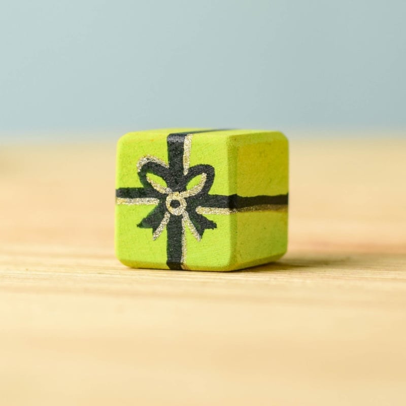 The Bumbu Handmade Wooden Christmas Present Set features a small green-painted wooden cube with a black ribbon and bow design, shown on a light wooden surface against a blue background—ideal for unique eco-friendly gifts.
