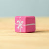 A small pink wooden "gift box" block from the Bumbu Handmade Wooden Christmas Present Set features a silver ribbon and bow design. Displayed on a light wood surface with a soft blue background—ideal for wooden gift enthusiasts.