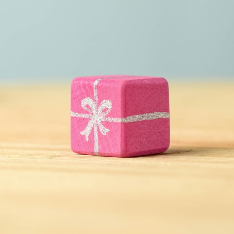 A small pink wooden "gift box" block from the Bumbu Handmade Wooden Christmas Present Set features a silver ribbon and bow design. Displayed on a light wood surface with a soft blue background—ideal for wooden gift enthusiasts.