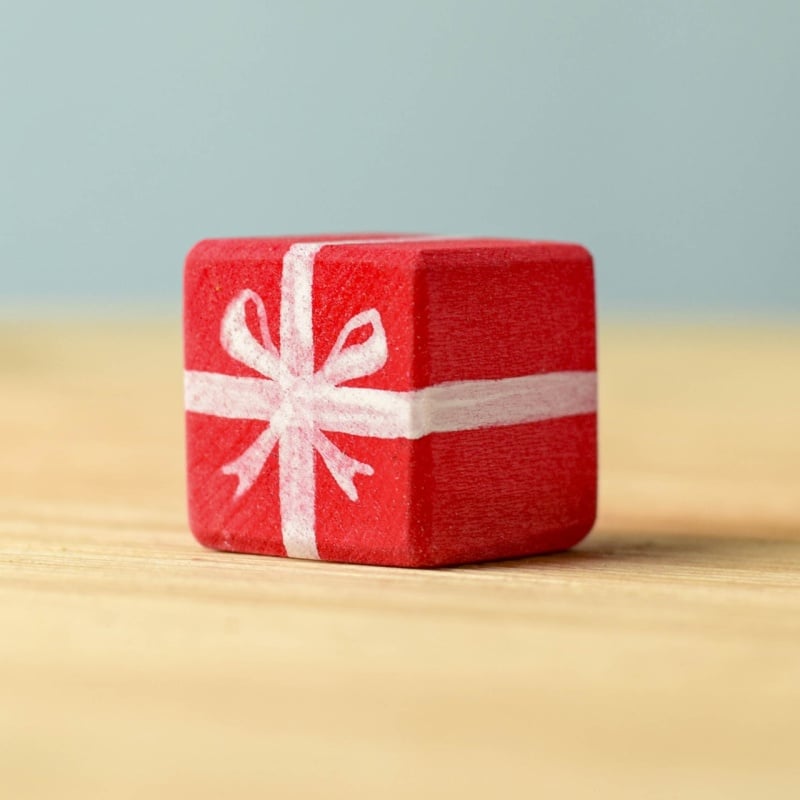The Bumbu Handmade Wooden Christmas Present Set features a small red gift box with a white ribbon and bow on a light wooden surface and soft blue background—an eco-friendly choice ideal for holiday gifting.
