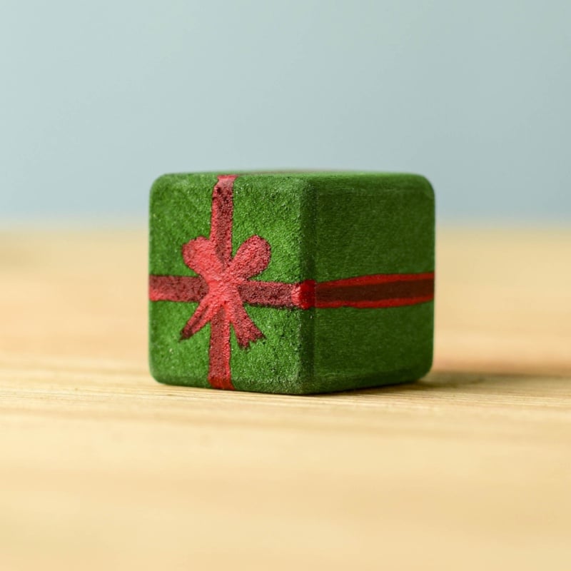 A small green cube gift box from the Bumbu Handmade Wooden Christmas Present Set, decorated with a red ribbon and bow, rests on a light wooden surface against a pale blue background.