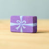 The Bumbu Handmade Wooden Christmas Present Set features a small, rectangular, eco-friendly wooden block painted purple with a light blue ribbon and bow design, ideal as a wooden gift. Shown on a light wooden surface against a pale blue background.