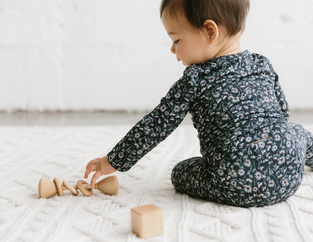 A toddler in a floral onesie plays with a Wooden Baby Rattle on a white textured blanket, focused on picking it up.