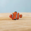 The Bumbu Handmade Wooden Clownfish, a small orange and white toy with black markings, sits on a light wood surface against a pale blue background—ideal for Montessori marine life learning.