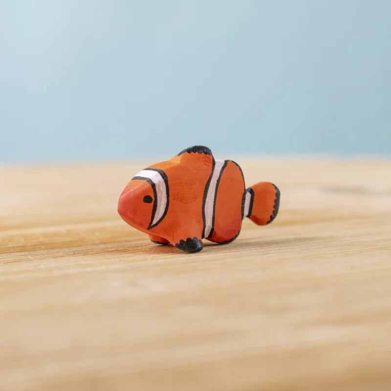 The Bumbu Handmade Wooden Clownfish, a small orange and white toy with black markings, sits on a light wood surface against a pale blue background—ideal for Montessori marine life learning.