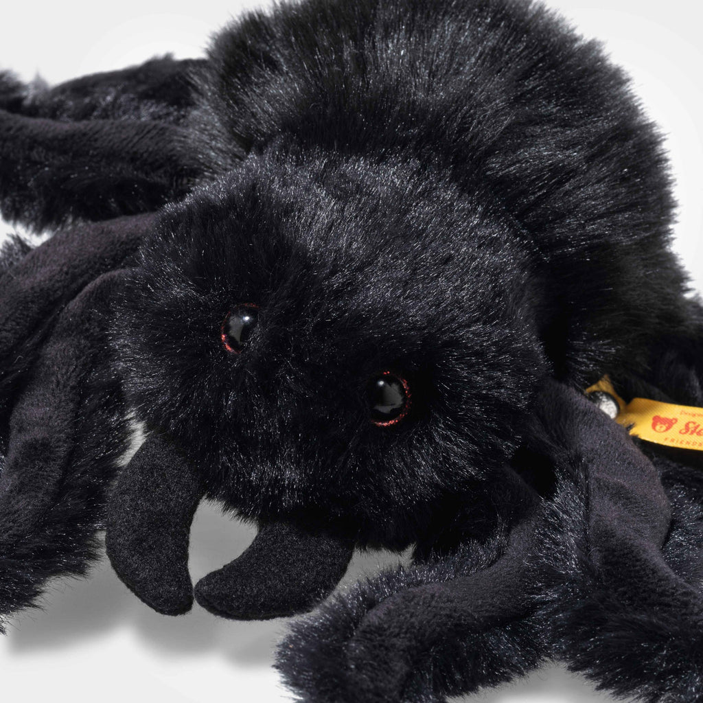 A close-up of Steiff Cobweb the Spooky Spider shows shiny black eyes, soft fuzzy fur, curved legs, and a yellow side tag marking it as an authentic stuffed animal.