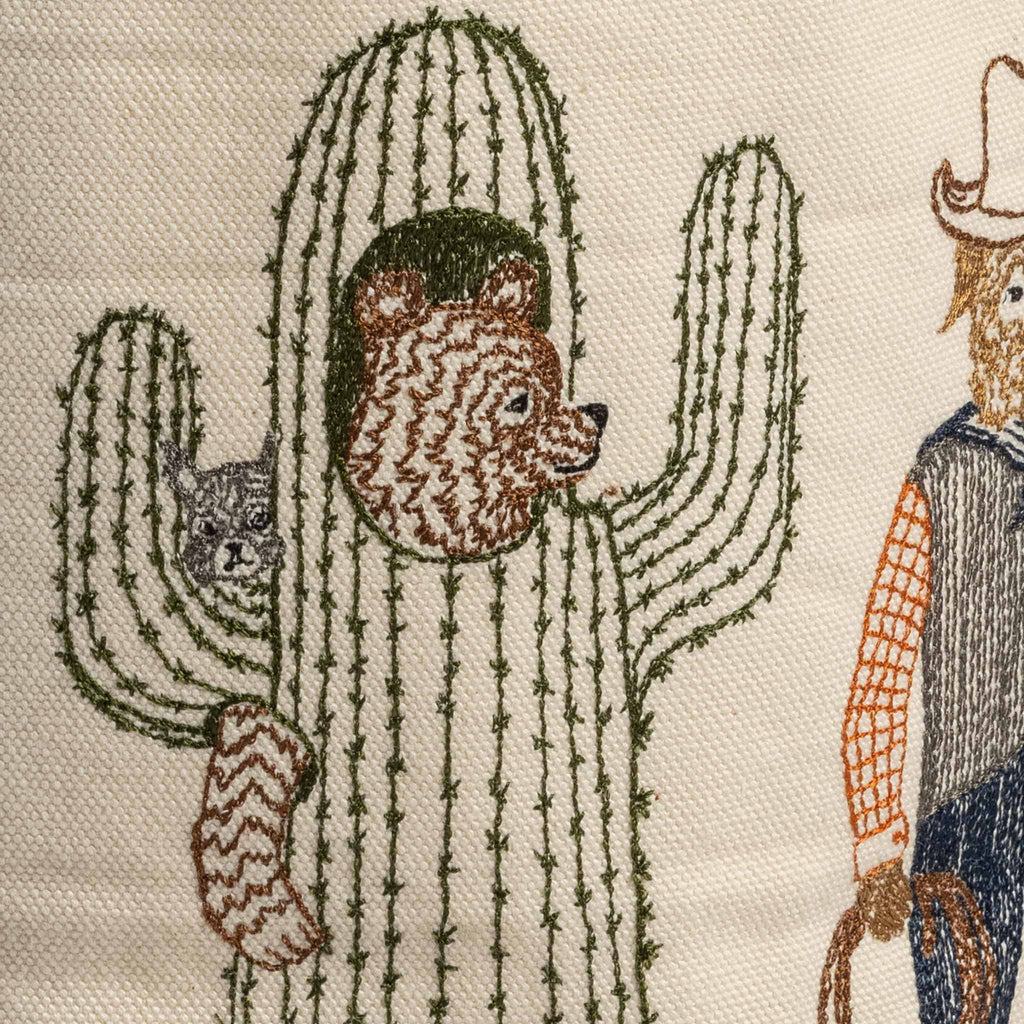 Embroidery of a whimsical scene featuring a bear peeking out from behind a large cactus, with a cowboy standing nearby on a beige fabric background, designed as an embroidered Trick or Treat Small Bucket cover.