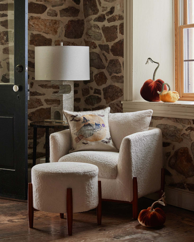 Cozy corner with a plush armchair, adorned with a Supermoon Spell Pillow. Next to a stone wall and window, seasonal pumpkins enhance the warm, autumnal ambiance.