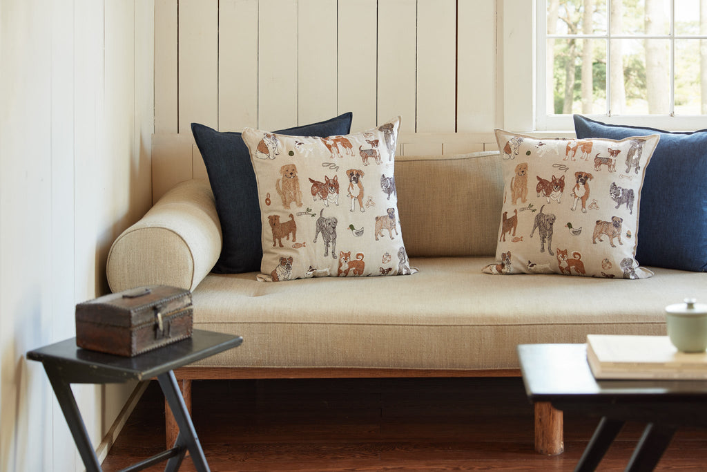 A cozy window seat with beige cushions, decorated with two blue and three Coral & Tusk Dogs and Toys throw pillows, next to a wooden side table with a small candle and an old book.