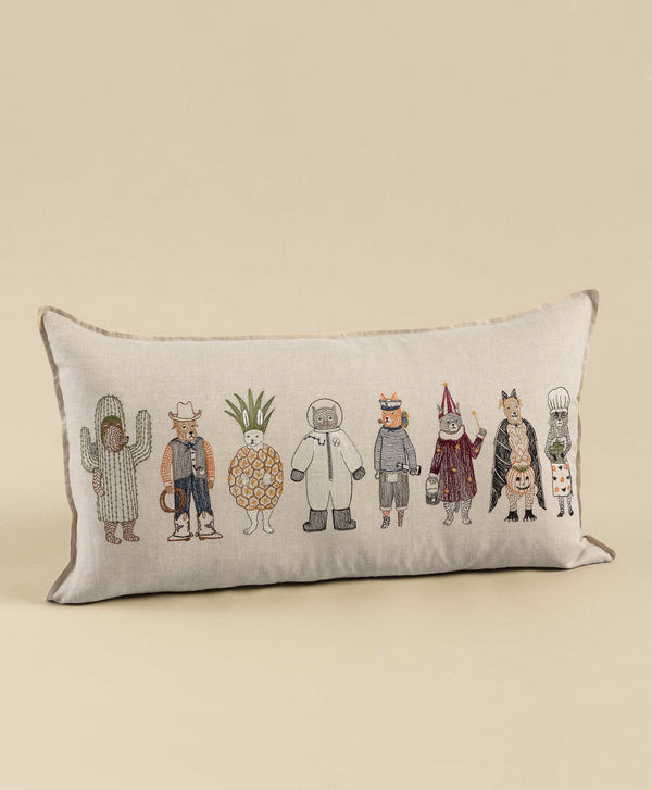 A Trick or Treat Pillow featuring a series of whimsical embroidered characters, including animals in Halloween costumes and a pineapple, on a neutral background.