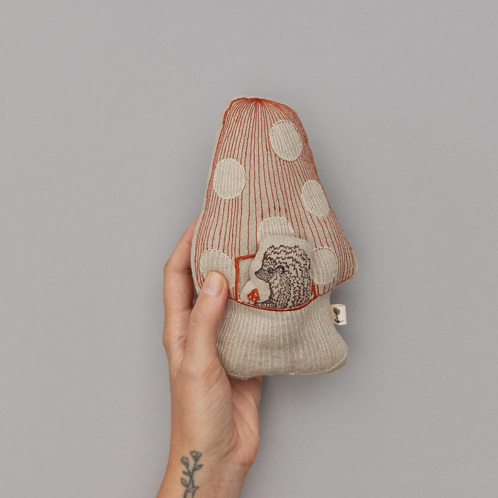 A hand with a floral tattoo holds a Coral & Tusk Pocket Mushroom decoration with red and white polka dots and intricately embroidered details against a gray background.