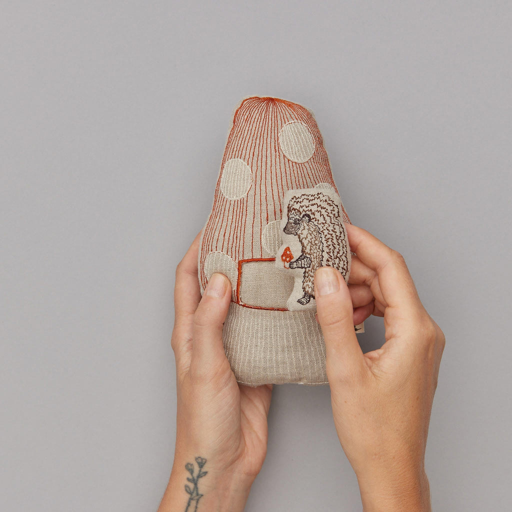 Sentence with product name: A person holding a Coral & Tusk Pocket Mushroom embroidered with a pattern of lines, dots, and a small figure, against a gray background. The person has a floral tattoo on their left wrist.