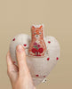 A hand holding a Coral & Tusk Fox Heart fabric pouch decorated with small red hearts, with an embroidered patch of a red cat holding a bouquet, against a beige background. This Coral & Tusk Fox Heart Pocket Valentine makes the perfect Valentine's Day.