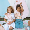 Two joyful children with curly hair, one holding an Olli Ella x Odin Parker Dinkum Dolls Cottontail Carrier – Hopscotch, celebrate Easter outdoors. They are surrounded by pastel eggs and decor; one child waves happily.