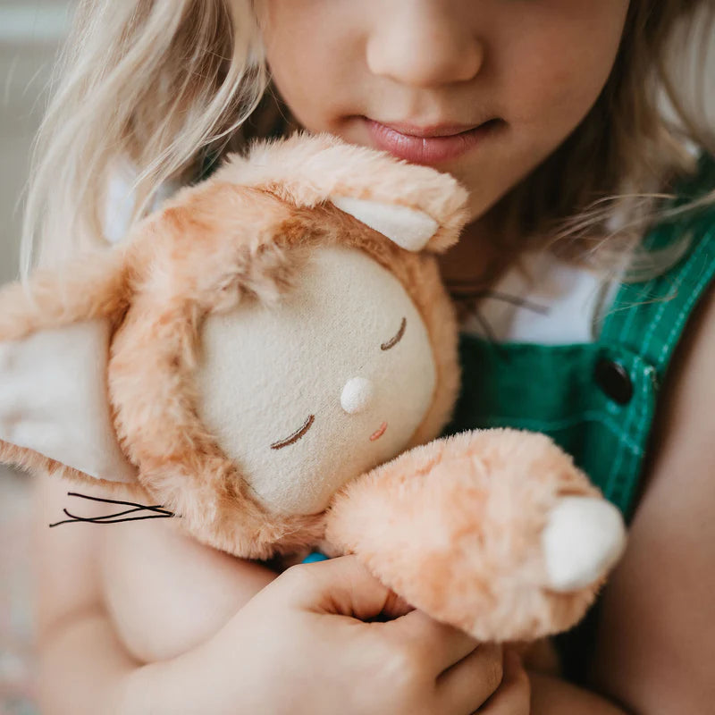 A serene child with blonde hair, dressed in a green outfit, gently holds an Olli Ella Cozy Dinkum Doll - Tabby Cat Jinx, which boasts a beige face and an orange-brown fuzzy body resembling an animal costume. The child is intently focused on the toy.