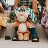 A child in green checkered pants and dark shoes sits outdoors, holding an Olli Ella Cozy Dinkum Doll - Tabby Cat Jinx. The toy, with its eyes closed, rests on the child's lap. Other children in colorful clothes are partially visible on either side.