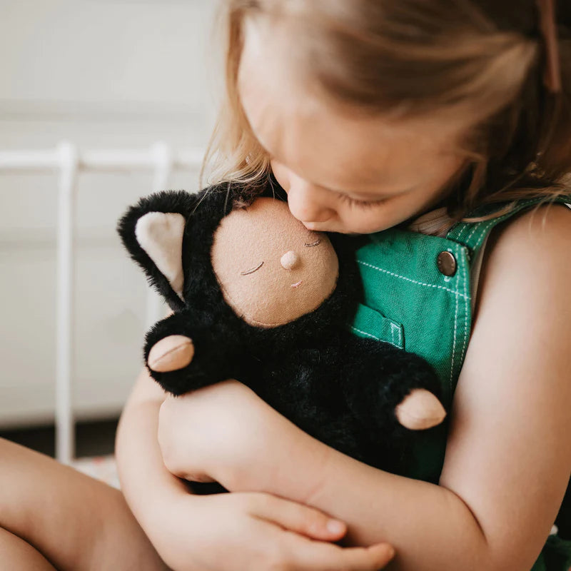 Sentence with product name: A young girl tenderly hugs her Olli Ella Cozy Dinkum Doll - Black Cat Nox, her cheek pressed against the doll's head. She is dressed in a cozy plush onesie and radiates warmth and comfort.