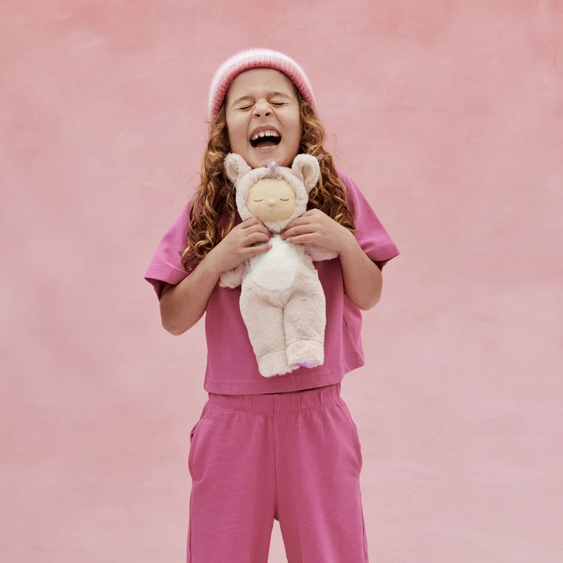 A child with long curly hair, wearing a pink beanie and outfit, laughs while holding the Olli Ella Cozy Dinkum Doll Unicorn - Sugarpie, featuring shimmering wings, against a pink background.