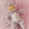 The Olli Ella Cozy Dinkum Doll Unicorn - Sweetpea, with a fluffy gray body, soft lavender belly fur, and a shimmering horn, rests on a pink surface amid scattered silver star confetti.