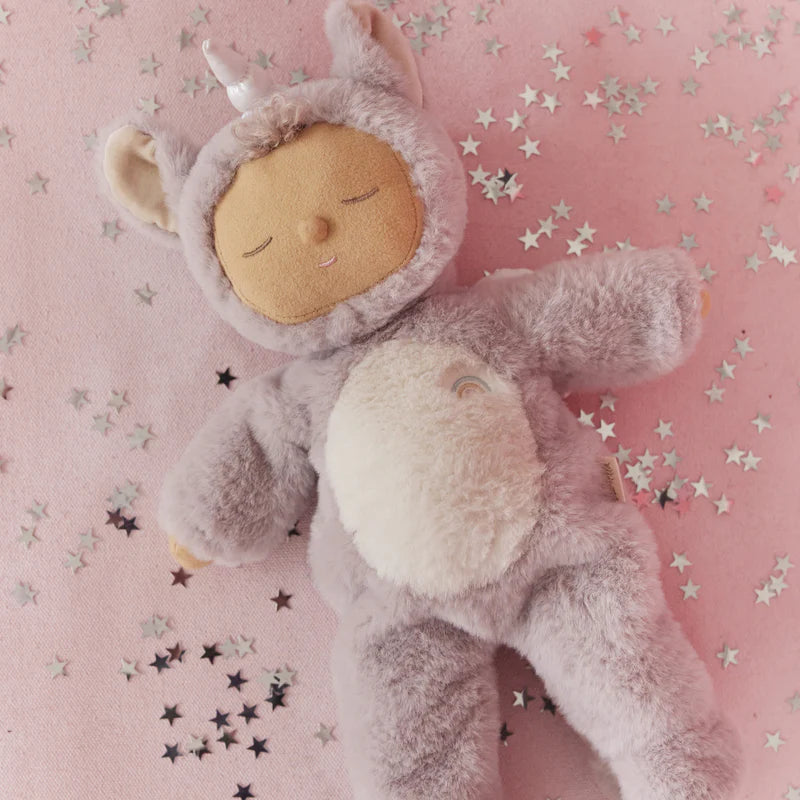 The Olli Ella Cozy Dinkum Doll Unicorn - Sweetpea, with a fluffy gray body, soft lavender belly fur, and a shimmering horn, rests on a pink surface amid scattered silver star confetti.