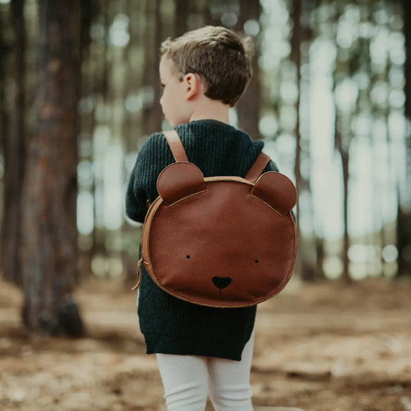 A young child with short hair, wearing a dark sweater and white tights, stands in a forest. The child has a Donsje School Leather Backpack - Bear on their back, facing away from the camera.