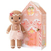 A hand-knit Cuddle + Kind Tiny Gingerbread - Sugarplum doll wearing a pink apron and bow, standing next to a house-shaped cardboard gift box decorated with gingerbread-themed illustrations.