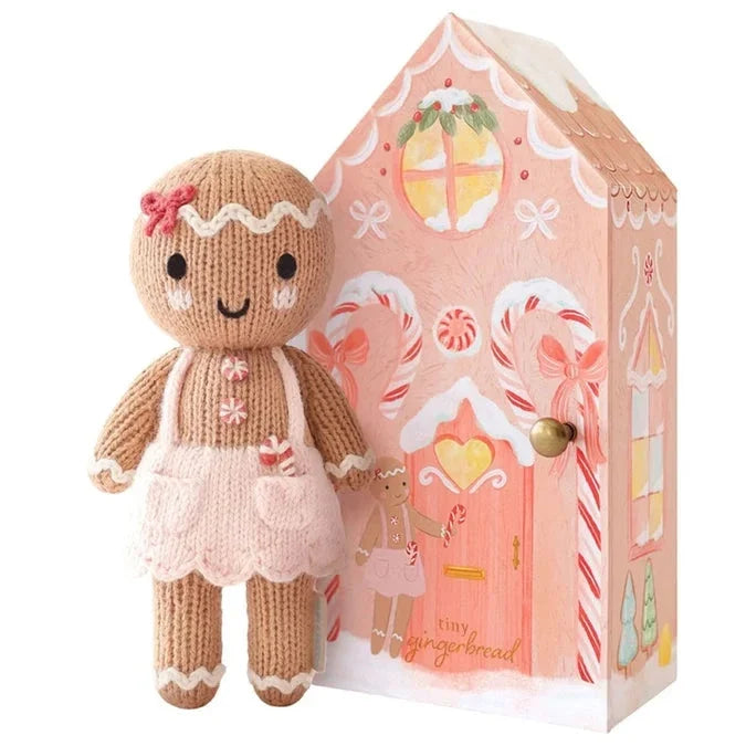 A hand-knit Cuddle + Kind Tiny Gingerbread - Sugarplum doll wearing a pink apron and bow, standing next to a house-shaped cardboard gift box decorated with gingerbread-themed illustrations.