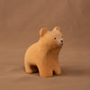 The Bumbu Handmade Wooden Curious Bear is a small wooden toy with simple painted facial features, displayed on a brown surface against a matching brown background.