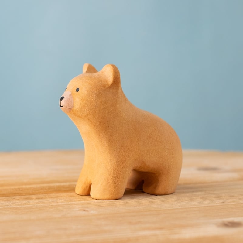 The Bumbu Handmade Wooden Curious Bear, a small light brown wooden toy, stands on a wooden surface against a plain light blue background—a delightful sensory development toy for toddlers.