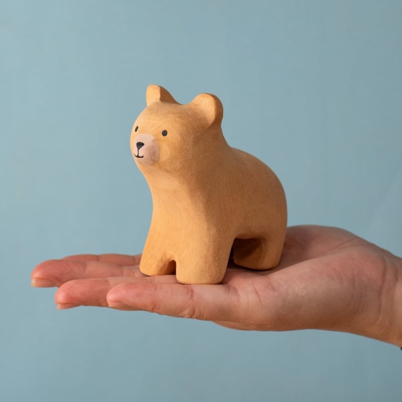A hand holds the Bumbu Handmade Wooden Curious Bear, a small light brown toddler wooden figure, against a plain light blue background.