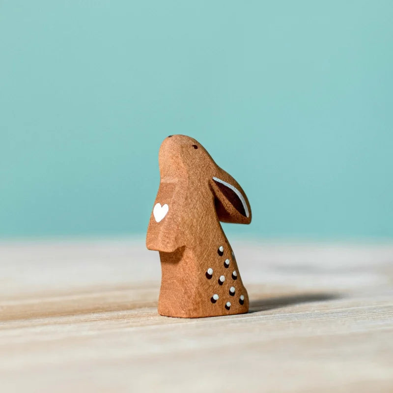 The Bumbu Handmade Wooden Curious Rabbit is a small, brown, handcrafted toy with a white heart and side dots. It stands upright on a light wooden surface against a blue background—an eco-friendly choice for play or Easter baskets.