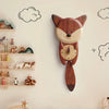 The Handmade Wooden Fox Wall Clock hangs on a light wall with cloud and swirl designs, bringing charm to the nursery decor. Animal figurines and toys on nearby shelves create a playful atmosphere.