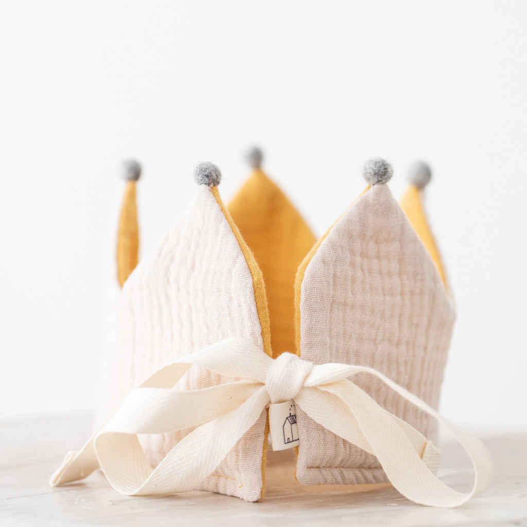 A handcrafted Reversible Crown made of yellow and beige organic cotton panels with gray pompoms on the tips, tied together with a beige ribbon featuring a small tag, set against a soft white background.