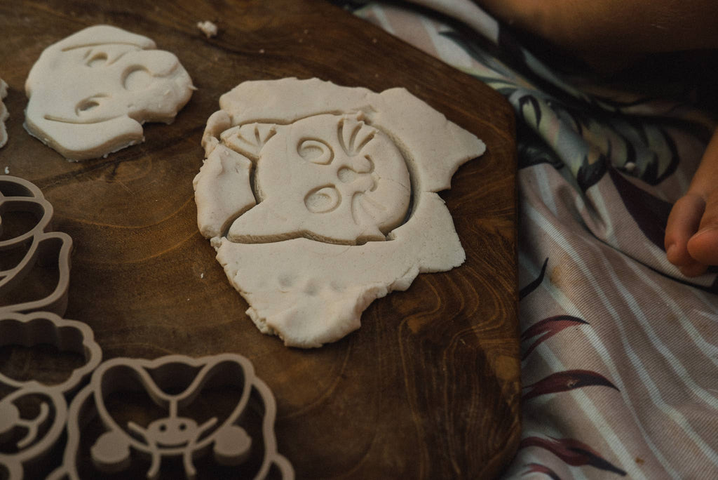 Cat-shaped dough is pressed and cut on a wooden surface, surrounded by more dough and the Pet Friends Faces Cutter Set - Eco Playdough Tool, with part of a person’s hand nearby. A striped cloth lies beneath the board, evoking the creativity and sensory play associated with using these eco-friendly playdough tools.