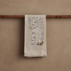 The Coral & Tusk Snowshoe Hare Tea Towel in beige linen hangs on a wooden rod, embroidered with a white rabbit beside a bare aspen tree for a serene winter scene.
